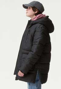 Giacca nera imbottita con cappuccio, dotata di zip frontale e tasche laterali. Indossata sopra un felpa bordeaux, abbinata a un cappello grigio.