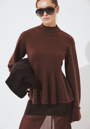 Vrouw draagt een donkerbruine top met lange mouwen, hoge hals en uitlopende zoom, draagt oversized zonnebril en houdt een opgevouwen zwart kledingstuk vast.