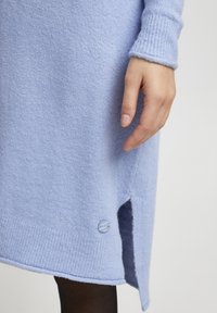 Un vestito a maglione di colore azzurro chiaro con orlo a coste, spacco laterale e un solo bottone decorativo vicino all'orlo. Tessuto morbido e maniche aderenti.