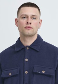 Camisa de forro polar azul marino con cierre de botones, dos bolsillos frontales y un cuello. Presenta botones marrones y una tela texturizada.