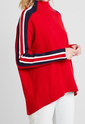 Rød oversized sweater med marineblå, hvide og røde striber langs ærmerne båret over hvide bukser på en person, der ses fra hals til lår.