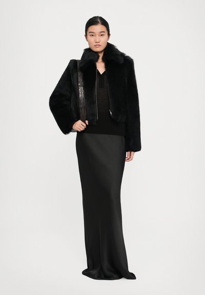 Woman wearing a black faux fur jacket, sheer black top, long black satin skirt, and carrying a dark brown textured shoulder bag.