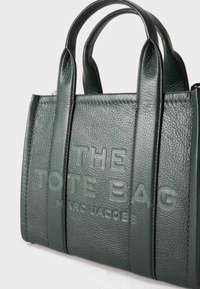 Sac cabas en cuir texturé vert foncé avec deux poignées et texte embossé "THE TOTE BAG MARC JACOBS" sur le devant.