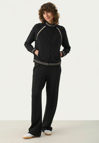 Svart idrottsjacka med hög krage och dragkedja, med kontrasterande beigea ränder. Kombinerad med svarta vida byxor och vita sneakers.