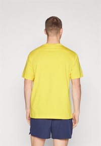 Camiseta de algodón amarilla con mangas cortas, cuello redondo y un corte relajado, mostrada desde atrás sobre un fondo blanco liso.