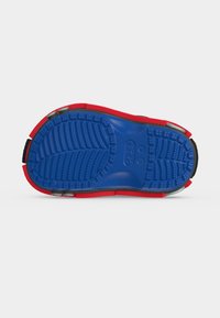 Σόλα παιδικού παπουτσιού Crocs κόκκινου και μπλε χρώματος με υφή αντιολισθητικής επιφάνειας και ορατό λογότυπο της μάρκας σε λευκό φόντο.