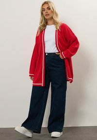 Röd ribbad cardigan med vit kant, ihop med en vit t-shirt och mörkblå vida jeans. Modell bär vita sneakers.