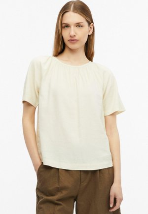 Jeune femme aux cheveux châtain clair et raides, portant une blouse crème à manches courtes et un pantalon marron, debout avec une main dans la poche.