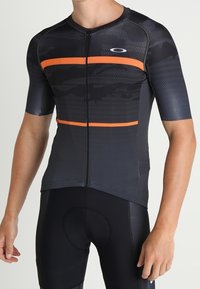 Lyhythihainen pyöräilypaita mustana, jossa on oranssit vaakaraitat. Liukas, joustava kangas vetoketjulla ja tekstuurikuvioiduilla yksityiskohdilla.