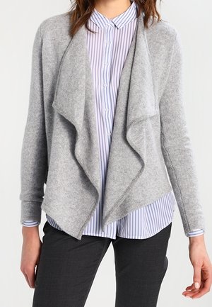 Kvinde iført en lys grå åben, draperet cardigan over en hvid og blå stribet skjorte med knapper og sorte bukser.