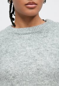 Hellgrauer Strickpullover mit gerippter Struktur, ausgestattet mit einem Rundhalsausschnitt und sichtbaren Steppnähten. Keine Muster oder Verzierungen vorhanden.