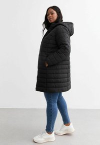 Abrigo de plumas negro con capucha, con textura acolchada vertical, cierre con cremallera y bolsillos laterales, combinado con vaqueros azules y zapatillas blancas.