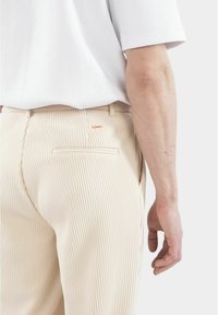 Beige randiga byxor med en slät textur, med en bakficka och en liten broderad logo i orange ovanför den högra fickan.