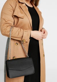 Borsa nera textured con tracolla a catena, abbinata a un trench beige con bottoni gialli e un pannello a motivi.