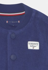Granatowy sweter robiony na drutach z okrągłym kołnierzem, dwoma dopasowanymi guzikami i białą metką z logo "tommy hilfiger".