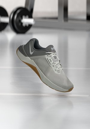 Zapatilla deportiva gris y blanca con suela de goma flotando en un gimnasio con mancuernas desenfocadas al fondo.