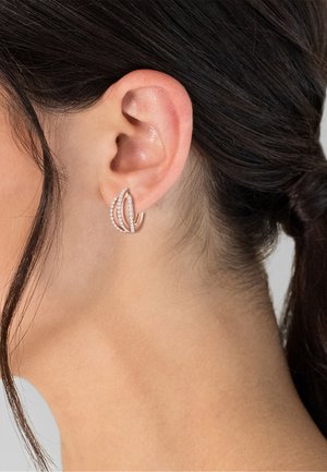 Kvinders øre med mørkt hår trukket tilbage, iført rosaguldfarvede trekoblede øreringe med små hvide sten på to af øreringene.