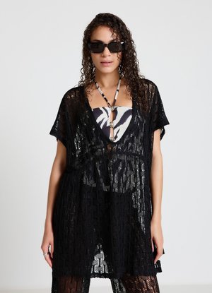 Donna con capelli ricci e occhiali da sole che indossa un copri abito in pizzo nero trasparente sopra un top a collo halter fantasia.
