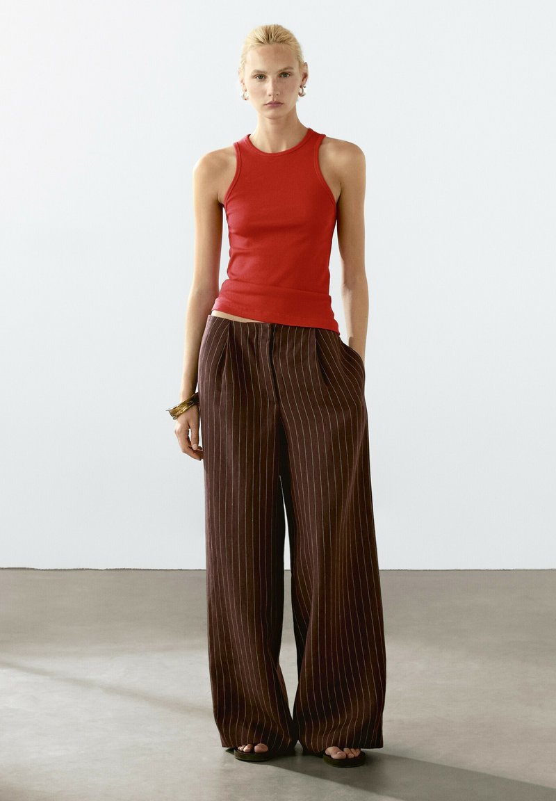 Rød ærmeløs bluse med en tætsiddende pasform, kombineret med løse brune pinstripede bukser med brede ben og frontfolder.