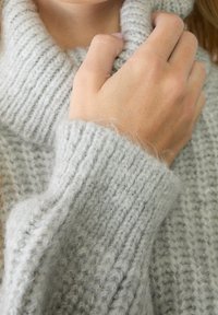 Main tirant le col d'un pull tricoté épais gris clair doux près du cou, mettant en valeur la texture et la chaleur.