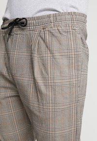 Ruudulliset olohousut harmaassa, mustassa ja oranssissa värimaailmassa. Varustettu joustavalla vyötäröllä, johon kuuluu nauha, sekä sivutaskuilla.