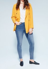Blazer amarillo de doble botonadura sobre una camiseta blanca de cuello en V, combinado con jeans azules ajustados y mocasines negros. Texturas suaves y diseño moderno.