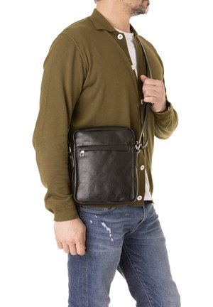 Zwarte leren crossbody tas met een gladde textuur, rechthoekige vorm, voorzak met ritssluiting en verstelbare schouderband. Gediend door een persoon in casual kleding.