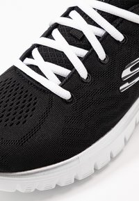 Chaussure de sport noire avec un dessus en tissu texturé, lacets blancs et semelle extérieure blanche. Présente des trous de ventilation et un logo de marque.