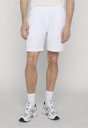 Man draagt witte sportshorts, witte sportsokken en witte New Balance sneakers, staand tegen een effen lichte achtergrond.