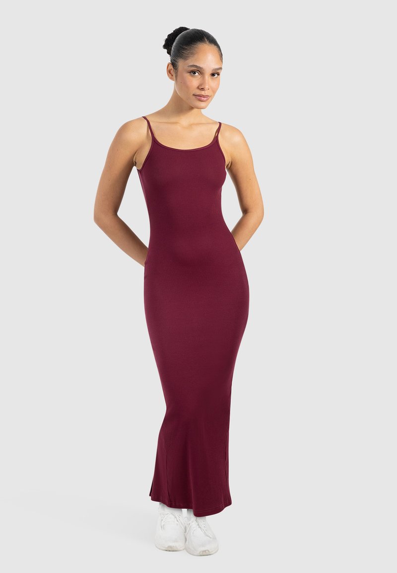 Burgunderfarbendes, geripptes Maxikleid mit dünnen Trägern, figurbetontem Schnitt und ausgestelltem Saum. Das Model trägt weiße Sneaker, der Stil wird vor einem hellen Hintergrund präsentiert.