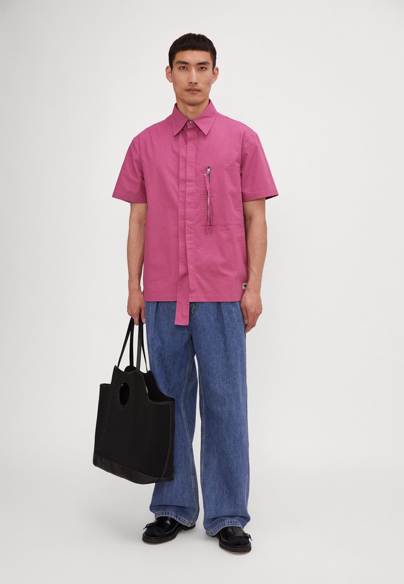 Camicia rosa a maniche corte con bottoni e tasca zip sul fronte, abbinata a pantaloni larghi in denim blu e una borsa a rete nera. Scarpe nere.