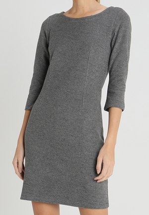 Vrouw draagt een nauwsluitende, knielange grijze jurk met driekwartmouwen en een ronde hals, staand tegen een effen achtergrond.