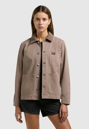 Giovane donna che indossa una giacca taupe con bottoni e quattro tasche frontali, abbinata a pantaloncini neri, in piedi davanti a uno sfondo chiaro e uniforme.