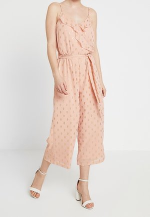 Femme portant une combinaison sans manches rose poudré avec un motif à pois dorés et un détail volanté, associée à des talons blancs à bride à la cheville.