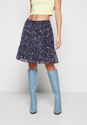 Kobieta w żółtym krótkim topie, fioletowej spódnicy w kwiatowy wzór i jasnoniebieskich kozakach do kolan, stojąca na tle jednolitej białej ściany.