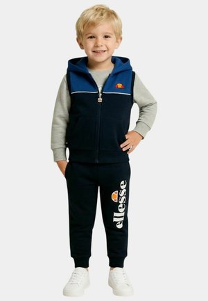 Garçon blond souriant portant une veste à capuche bleue, un t-shirt à manches longues gris, un pantalon noir avec le logo "ellesse" et des baskets blanches, debout la main sur la hanche.