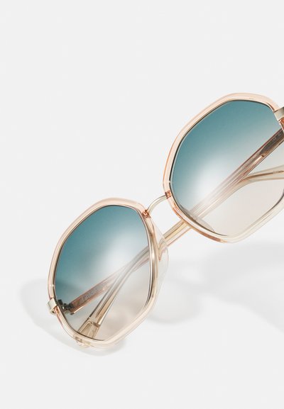 Occhiali da sole esagonali con montature beige traslucide e lenti sfumate blu appoggiati su una superficie bianca.