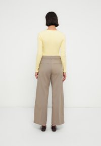 Γυναίκα με κοντά σκούρα μαλλιά, όρθια αντικρίζοντας έναν λευκό τοίχο, φοράει ανοιχτό κίτρινο μπλουζάκι με μακριά μανίκια, μπεζ παντελόνι φαρδύ, και σκούρα παπούτσια.