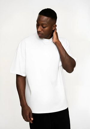 Hvid oversized t-shirt med korte ærmer og høj halsudskæring, lavet af glat stof. Modellen poserer med hovedet vippet, hånd på nakken.