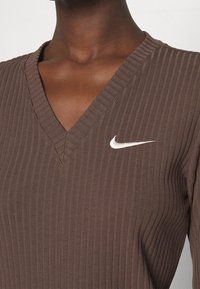 Haut marron à col en V côtelé en tissu doux, avec un logo Nike blanc sur le côté gauche de la poitrine. Texture détaillée visible.