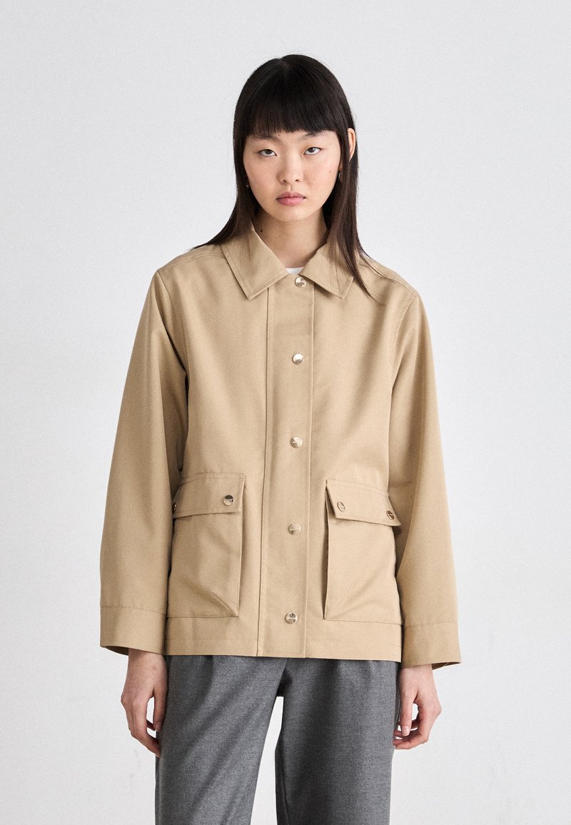 Beige utility jacket with a collared neckline, front buttons, and two chest pockets. Made of durable fabric with a loose fit and straight sleeves.
