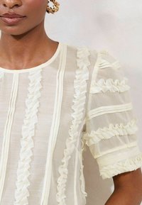 White, sheer blouse featuring vertical ruffles and horizontal stripes on short sleeves. Fabric has a light, textured appearance.