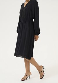 Robe noire à manches longues avec un col en V, des détails en dentelle délicate sur le corsage et l'ourlet, une taille élastique et une silhouette fluide.