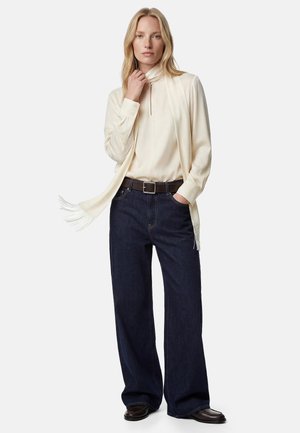 Kvinde iført cremefarvet rullekrave med frynset tørklæde, mørke vide jeans, brun bælte og mørkebrune sko, stående med hånd i lommen.