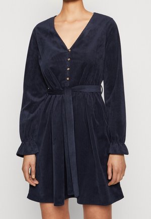 Woman wearing a navy blue, long-sleeve, corduroy dress with a V-neck, buttoned front, belted waist, and slightly ruffled cuffs.