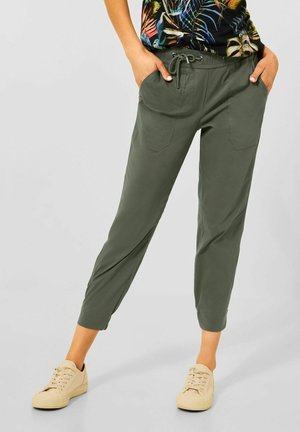 Personne portant un pantalon court vert olive avec des poches avant et des baskets beiges, debout les mains dans les poches.