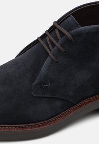 Navy plavutenice od suède s smešnim pertlama i detaljem ušivanja. Zaobljeni vrh i kontrastna smeđa potpetica s teksturiranom završnom obradom.