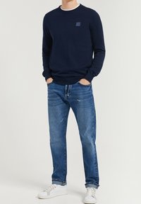 Man i mörkblå långärmad tröja, blå uppvikta jeans med mindre slitage och vita sneakers, står med händerna i fickorna.