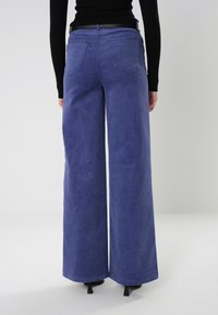 Pantalones de pierna ancha en corduroy morado con tejido texturizado, bolsillos traseros y una cintura alta, combinados con una blusa negra y tacones.