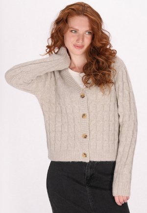Beige gebreide cardigan met kabelpatroon, v-hals, vijf houten knopen en geribbelde manchetten, gecombineerd met een zwarte rok.